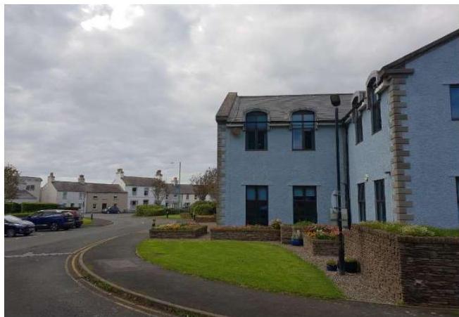 A street-level photograph showing a light blue building with stone detailing on the right, featuring low brick boundary walls and landscaping, with cars parked on the road to the left.
