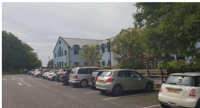 A photograph showing a row of parked cars in a lot with a large light blue building in the background.