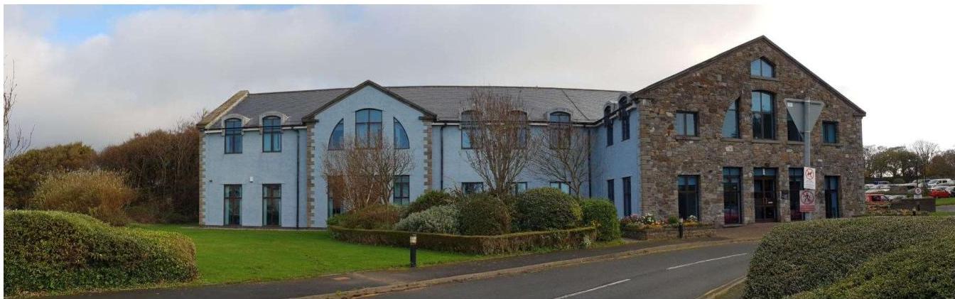 A panoramic photograph showing a large building complex with a mix of modern blue and older stone architecture, featuring foreground landscaping and a road.