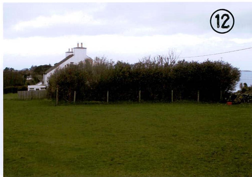 A photograph showing a grassy field in the foreground with a hedge and a white building in the background, with the sea visible to the right.