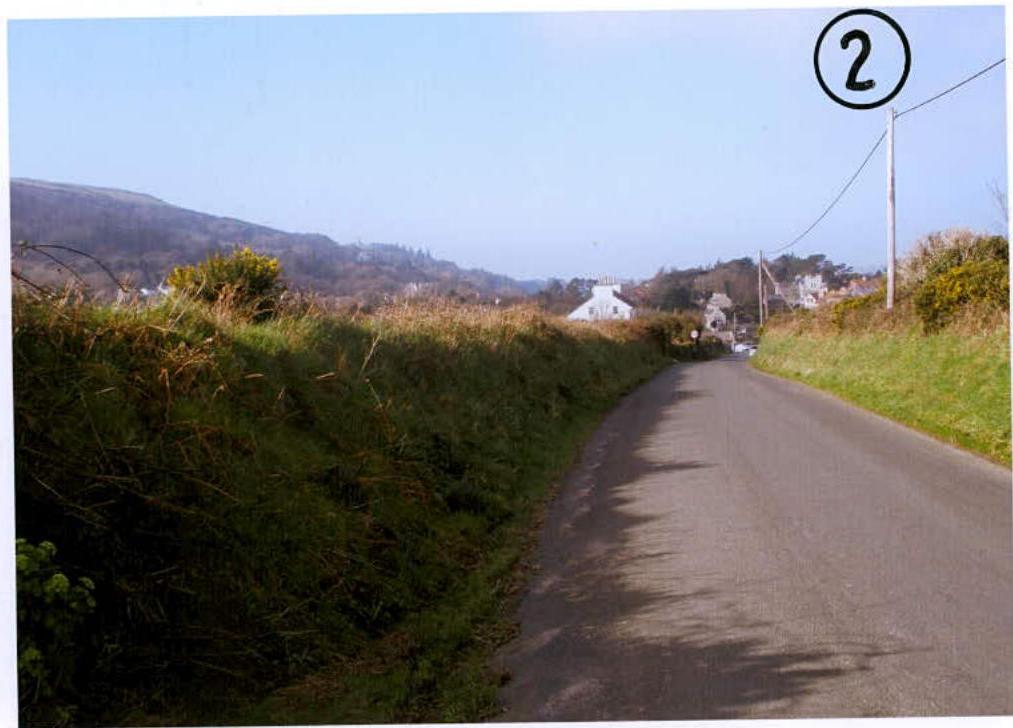 A photograph showing a rural road with grassy verges and hills in the background, likely depicting the site location.