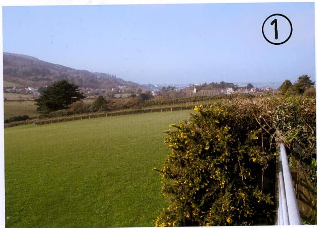 A photograph showing a large green agricultural field with hedgerows, overlooking a rural landscape and coastal town in the distance.