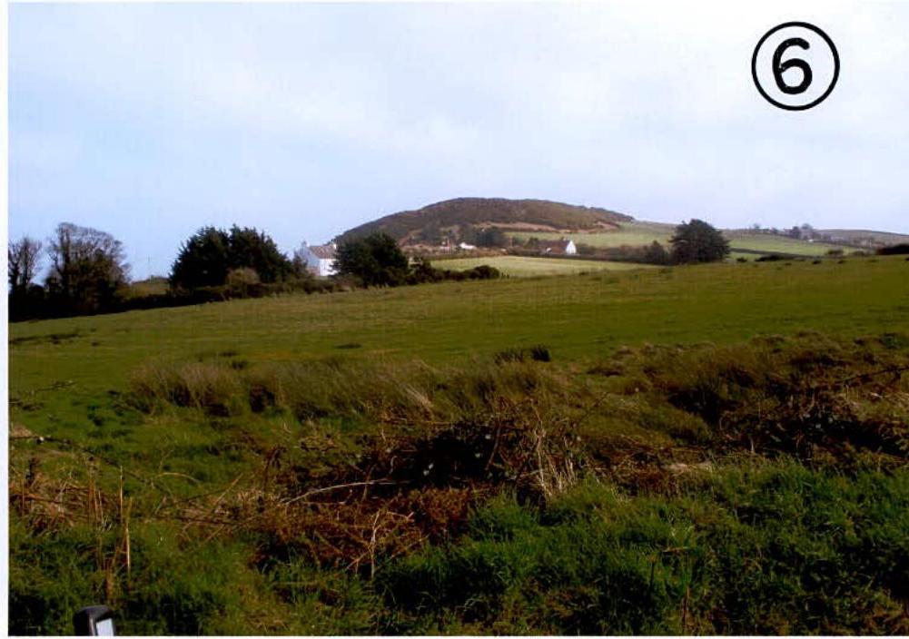 A photograph showing a green agricultural field in the foreground with a hill and scattered white houses in the background, illustrating the rural setting.