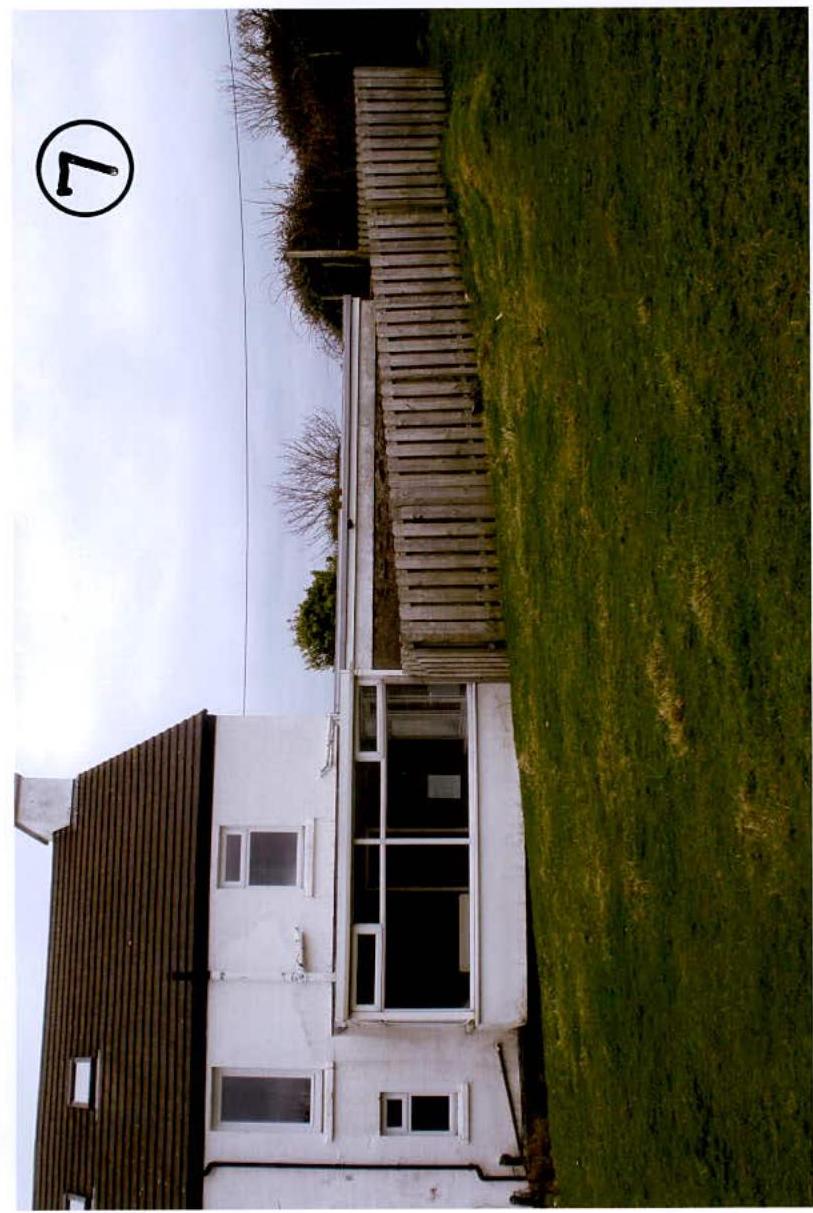 A rotated photograph showing a white building with a large glass extension adjacent to a wooden fence and grassy field.