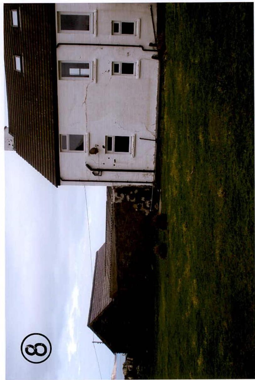 A rotated photograph showing a white two-story building adjacent to a grassy field and a smaller dark outbuilding.