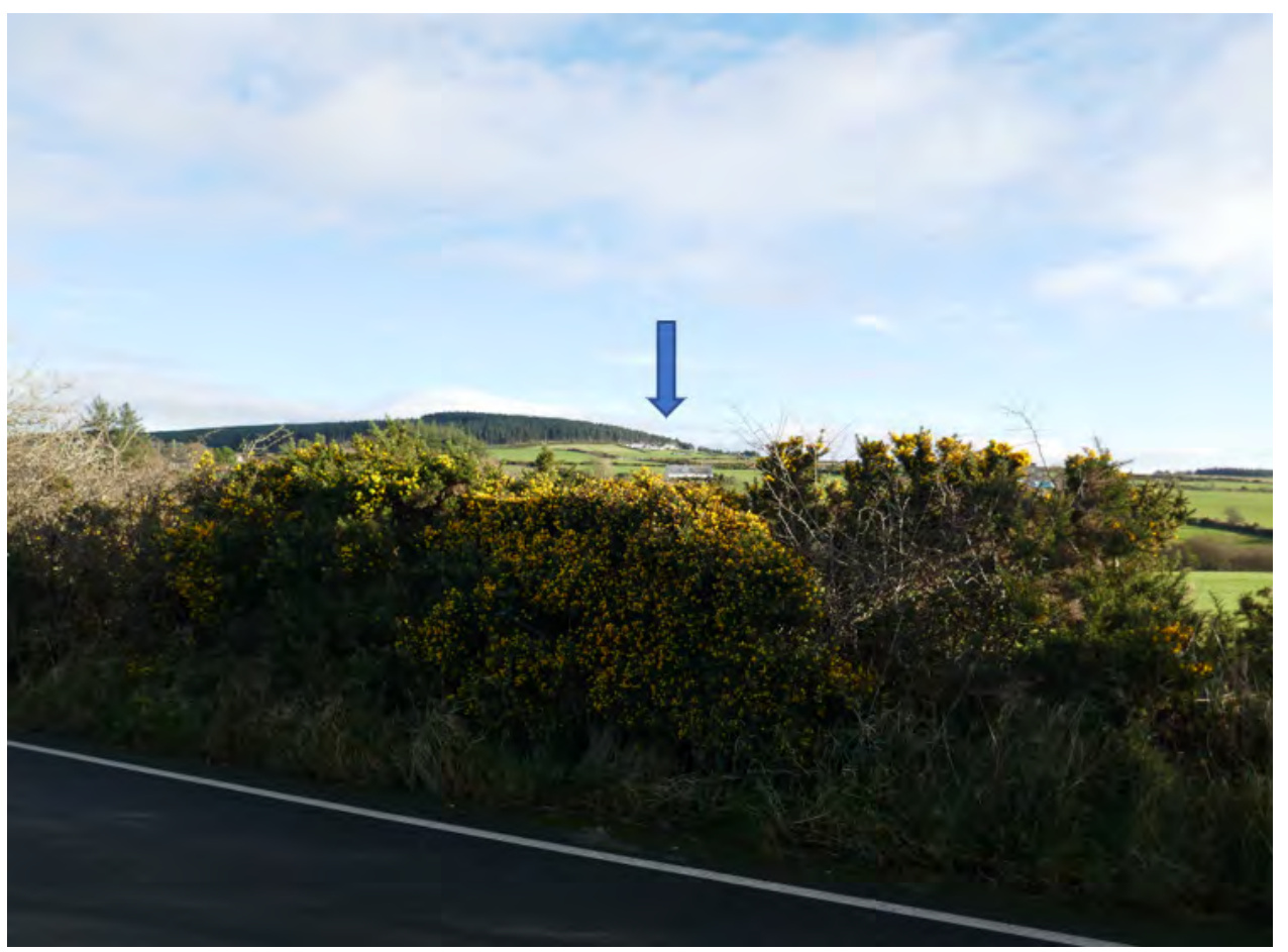 A landscape photograph showing a roadside verge with gorse bushes in the foreground and a blue arrow pointing to a distant site location in the rolling countryside.