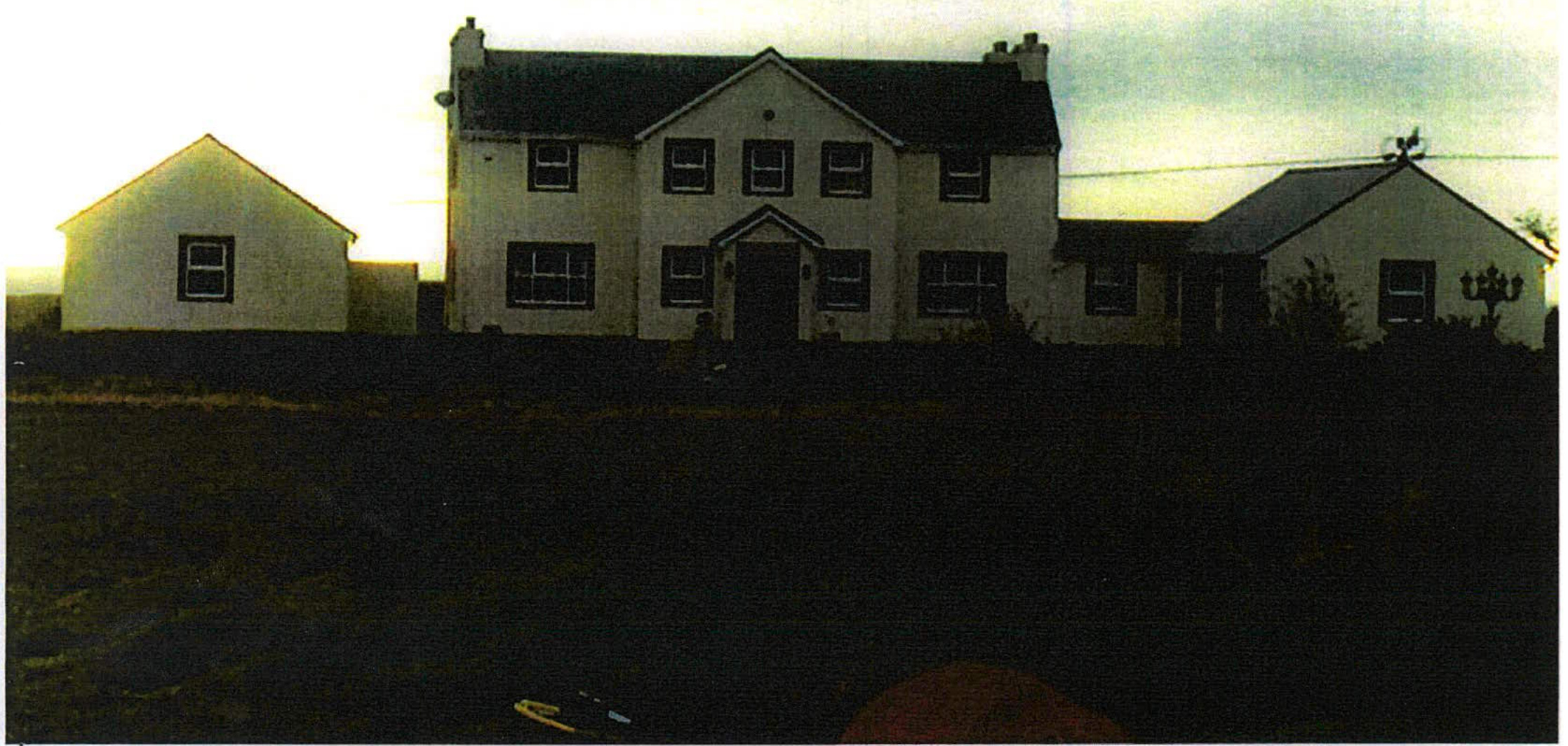 A grainy, low-light photograph of a large white two-story detached house with adjacent structures, likely showing the existing site or proposed replacement dwelling.
