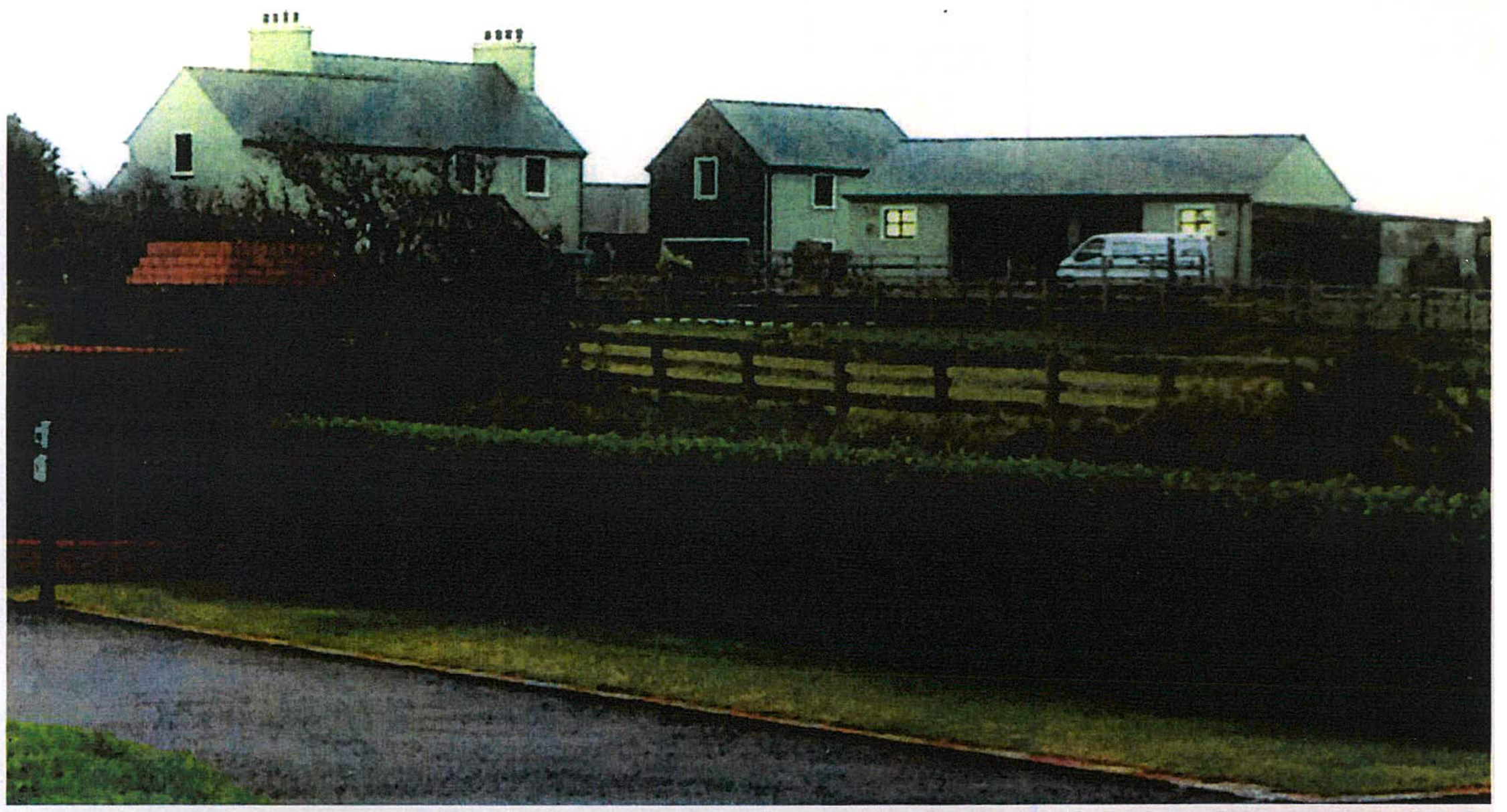 A photograph showing a rural property featuring a white detached house and an adjacent agricultural-style building with a wooden fence and hedge in the foreground.