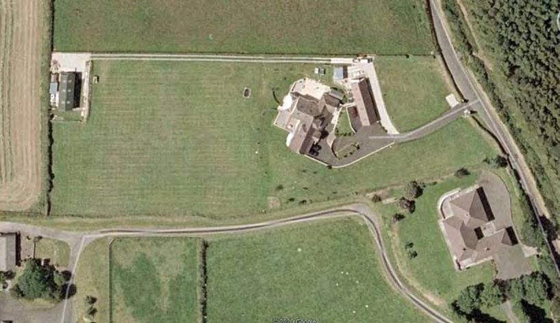 An aerial satellite view of a rural property plot showing existing buildings, green fields, and access roads.