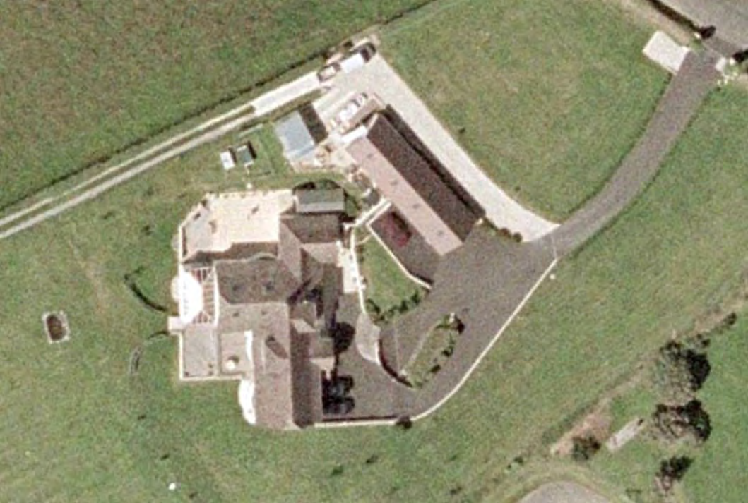 An aerial satellite view of a rural property featuring a large detached dwelling, driveway, and surrounding green fields.