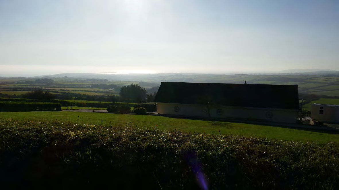 A photograph showing a large, single-story building situated on a grassy plot with a rural landscape and hills in the background. The image captures the existing site context for a proposed replacement dwelling.