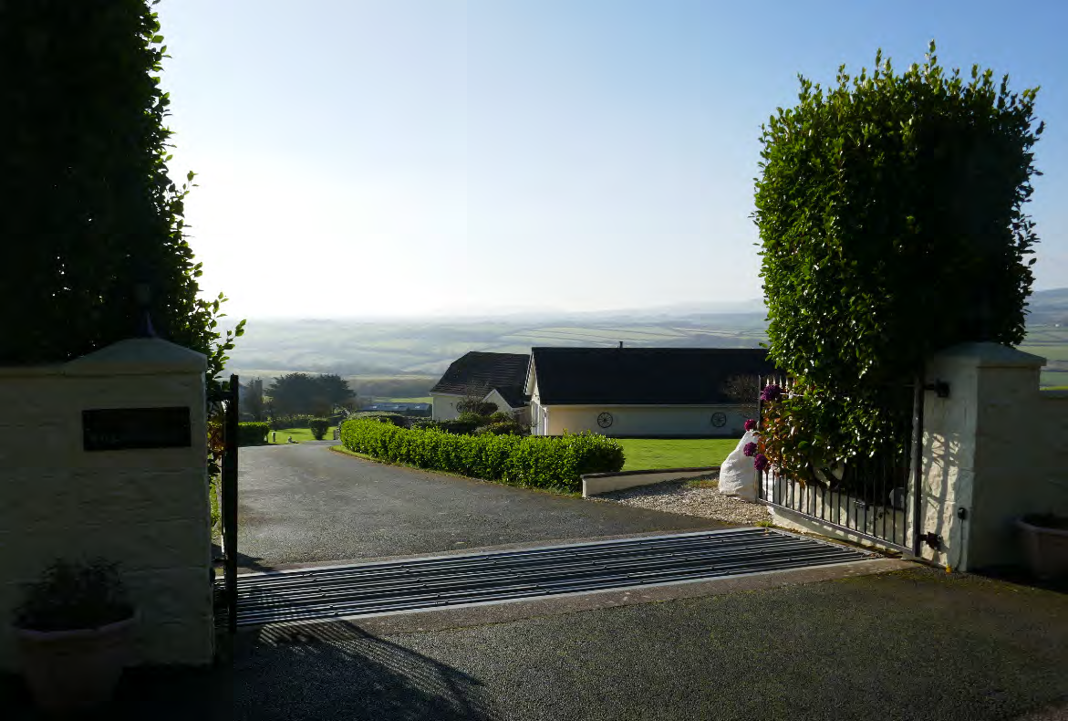 A photograph showing the property entrance with white pillars and gates, looking out over a driveway towards a detached white house and a wide rural coastal landscape.
