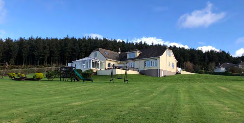 A photograph of a large detached house featuring a conservatory extension, set on a grassy slope with a dense forest background.