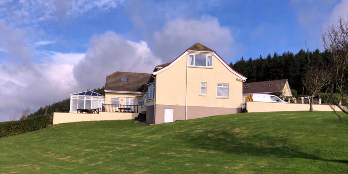 A photograph showing a large detached house on a grassy slope with a conservatory, likely the existing site for the proposed replacement dwelling.