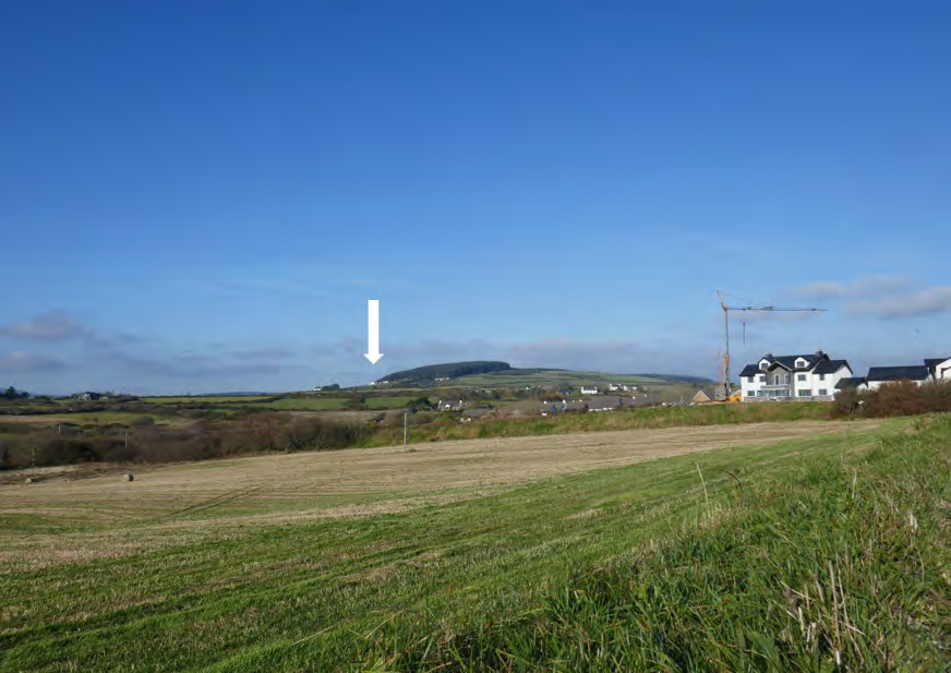 A landscape photograph showing a grassy field in the foreground with a white arrow pointing towards a distant hill, indicating the site location within a rural setting.