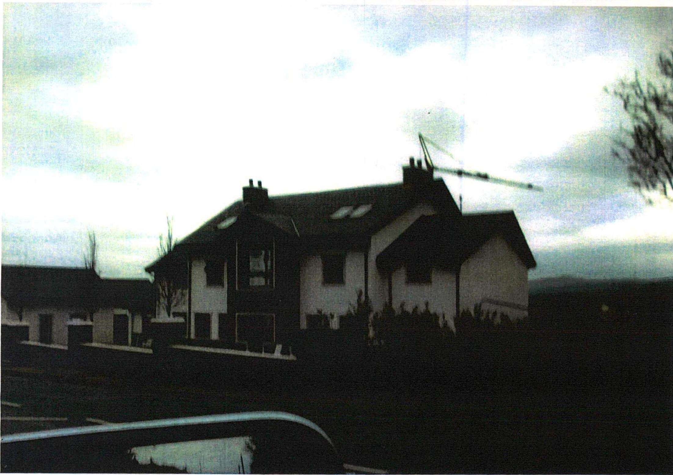 A grainy, low-light photograph showing a large, modern detached house with a dark roof and white walls. A construction crane is visible in the background against a cloudy sky.