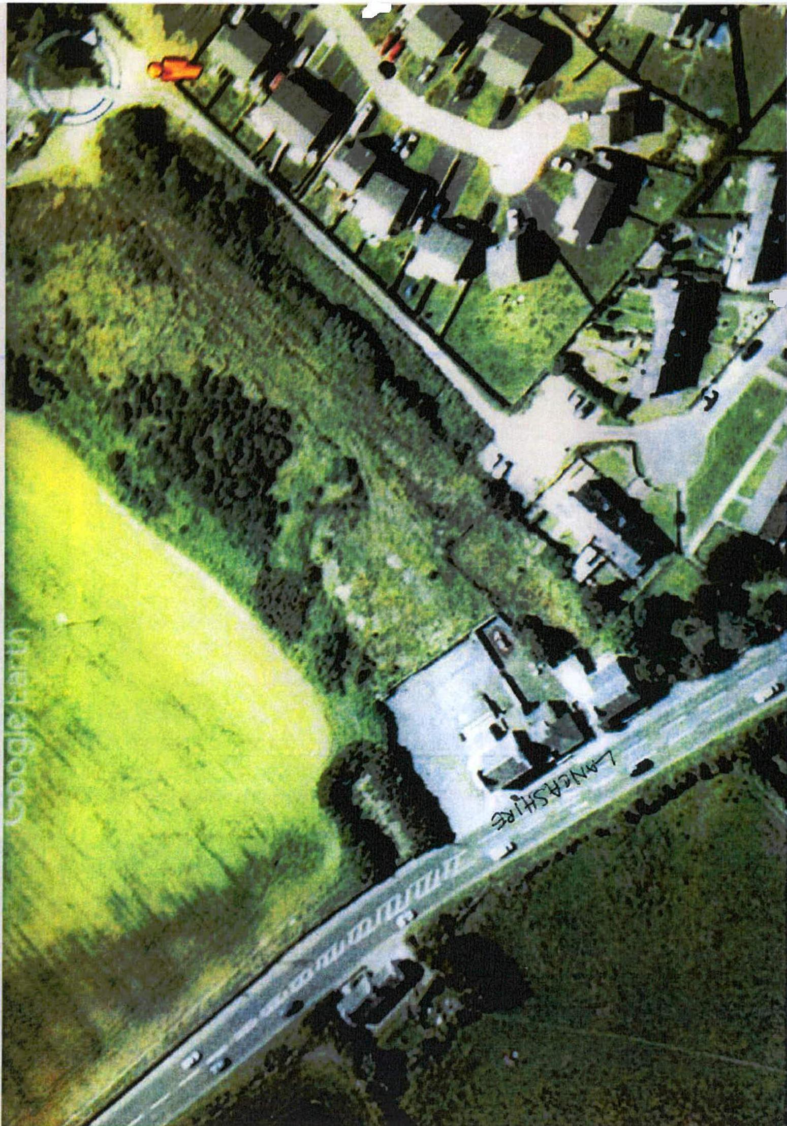 An aerial satellite view showing a road, surrounding residential properties, and a large green field, likely serving as a location plan for the planning application.