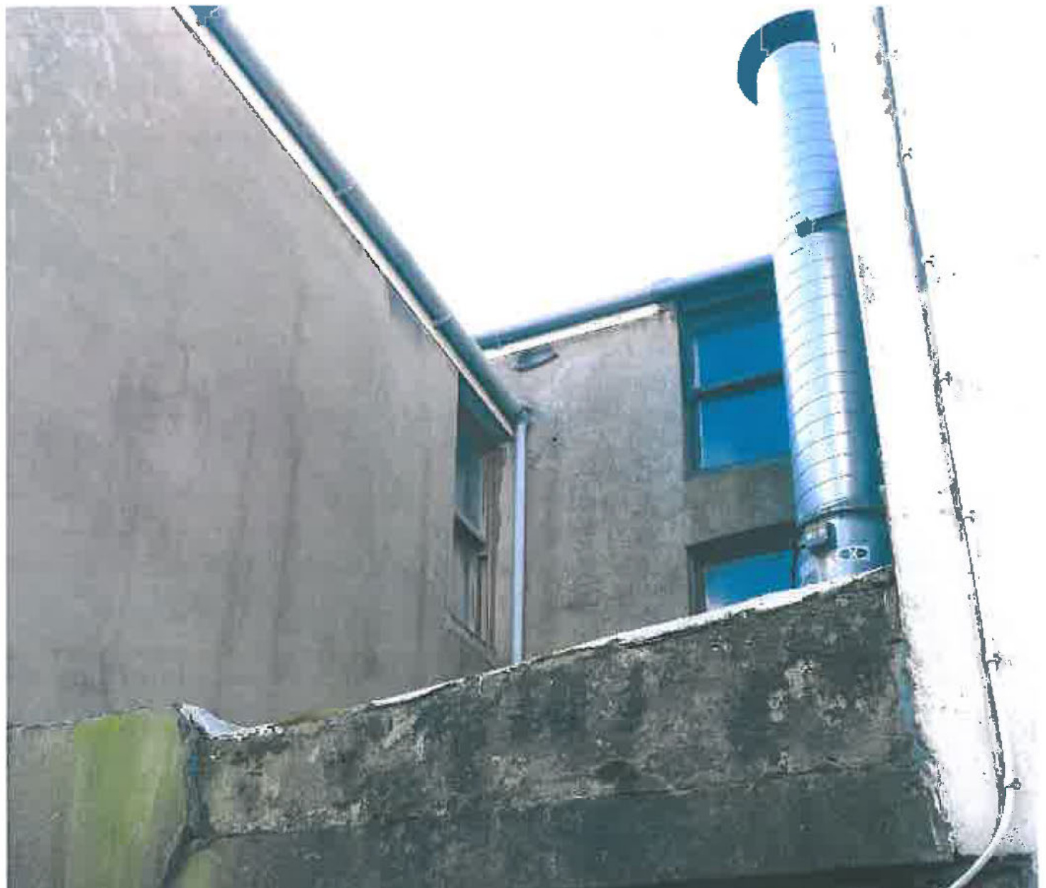 A close-up photograph showing a large silver metal extractor flue pipe attached to the side of a grey rendered building with blue windows.