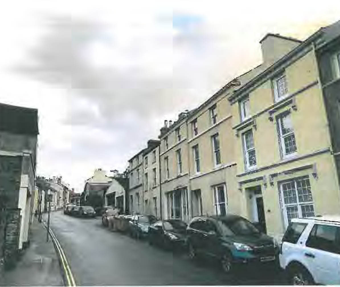 A street-level photograph showing a row of terraced residential buildings along a road with cars parked along the curb.