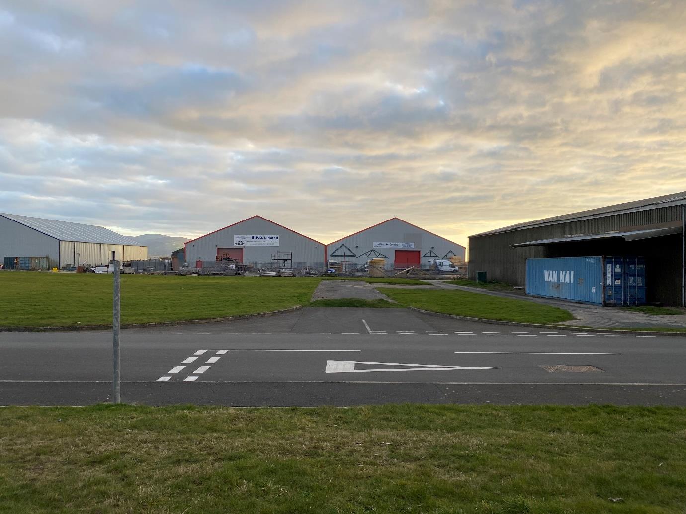 A street-level photograph showing a row of industrial warehouse units across the road, featuring a blue shipping container and road markings in the foreground.