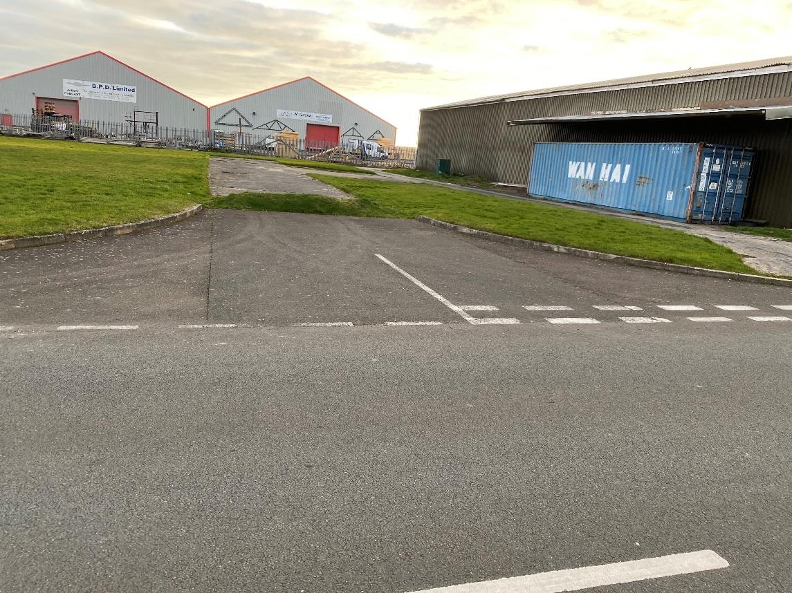 A street-level photograph showing an industrial site with warehouse buildings, a blue shipping container, and road access.