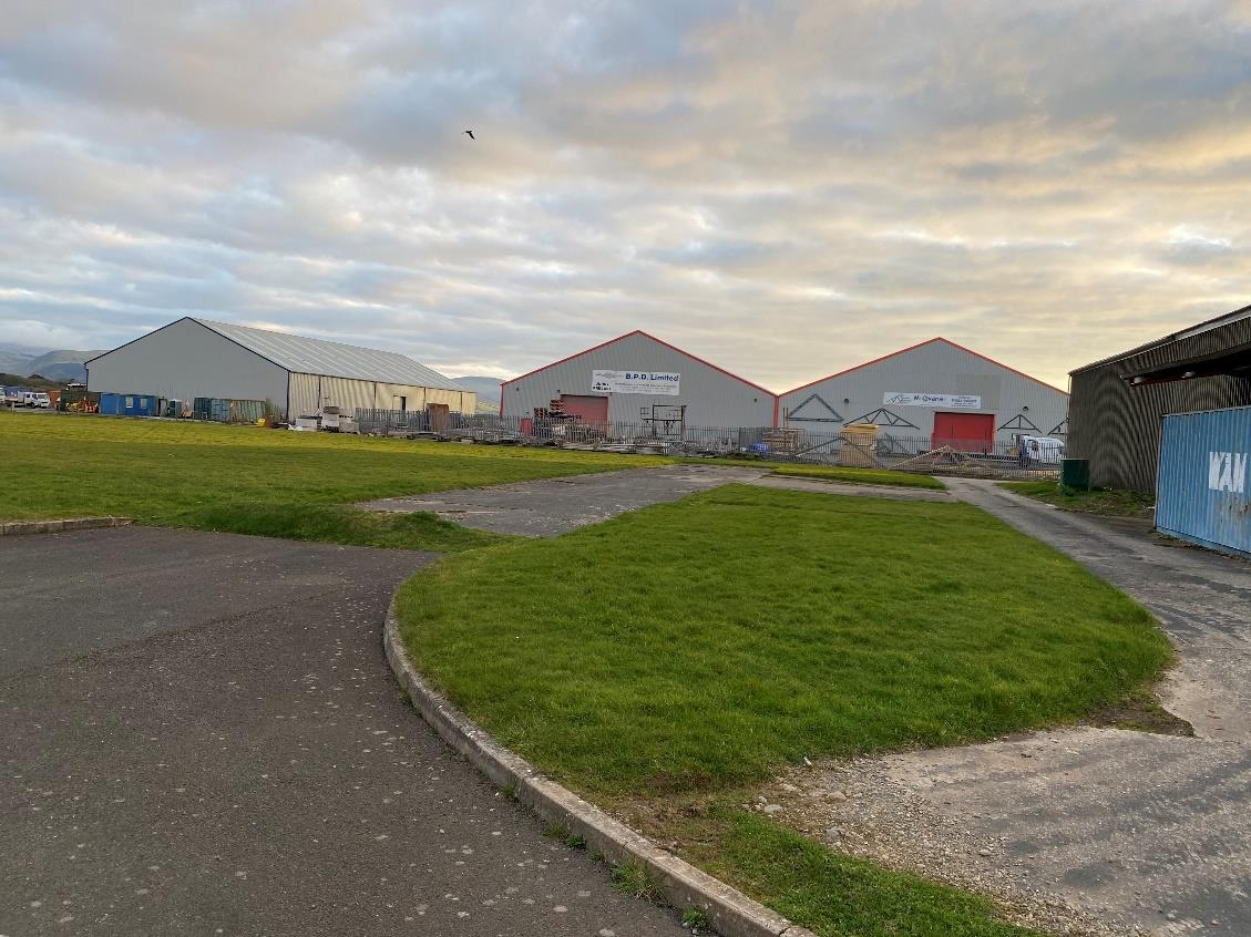 A photograph showing an industrial or commercial site with large warehouse buildings, paved access roads, and grassy areas under a cloudy sky.