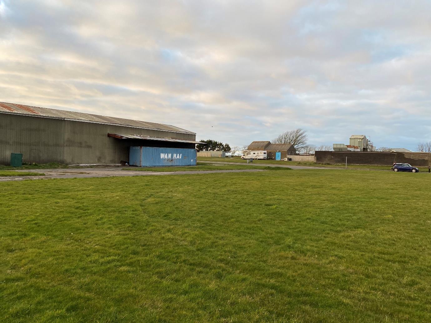 A photograph showing a large grassy field in the foreground with a corrugated metal industrial building and blue shipping container in the background.