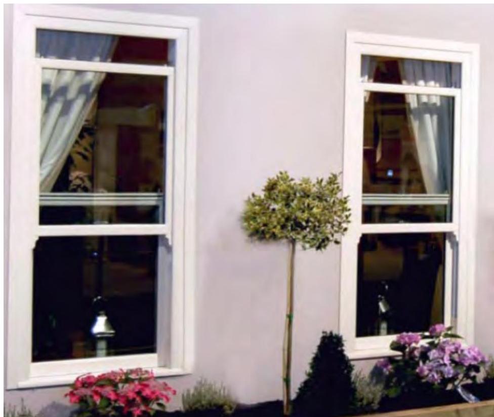 A photograph showing two white sliding sash windows installed in a light-colored wall, with garden plants and flowers in the foreground.