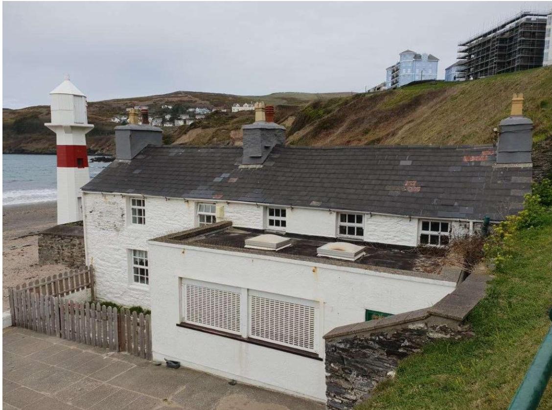 A photograph showing a white single-story building with a slate roof situated directly adjacent to a sandy beach and a small lighthouse.