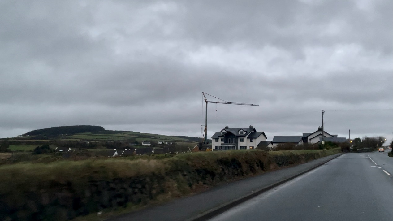 A roadside photograph showing a white detached house with a construction crane nearby, set against a rural landscape with rolling hills under a cloudy sky.