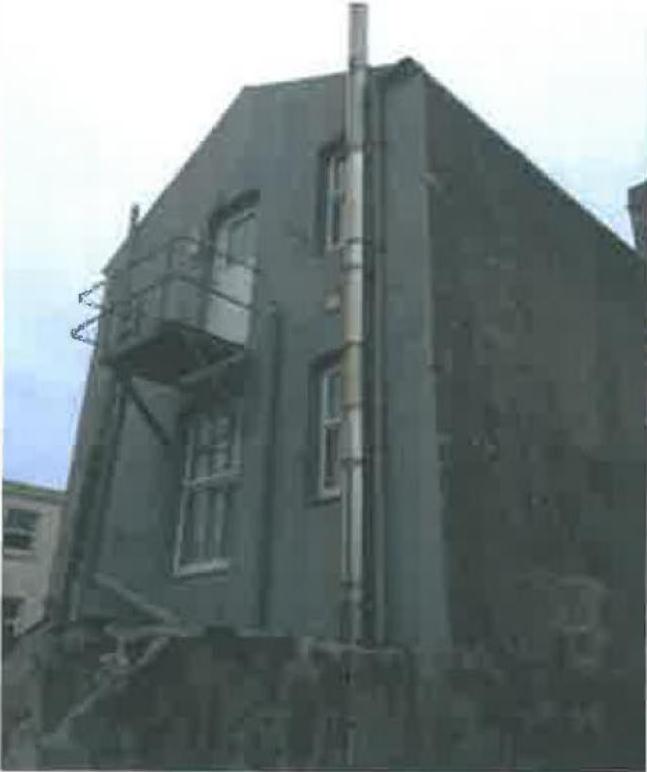 A low-angle photograph showing the side elevation of a two-story building with a prominent vertical flue pipe and a small balcony.