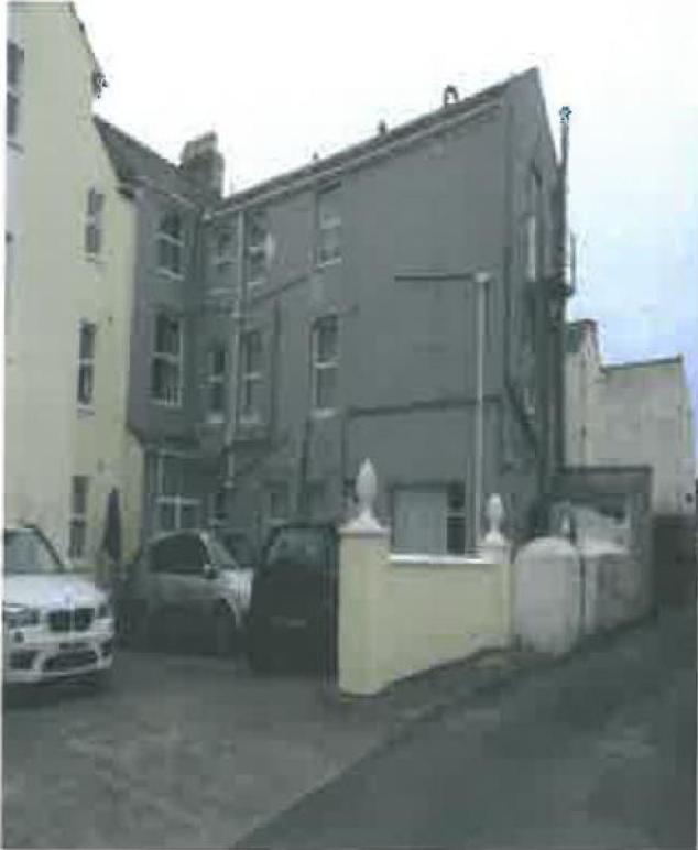 A grainy photograph showing the exterior of a grey, multi-story terraced building with cars parked in front and a low boundary wall.