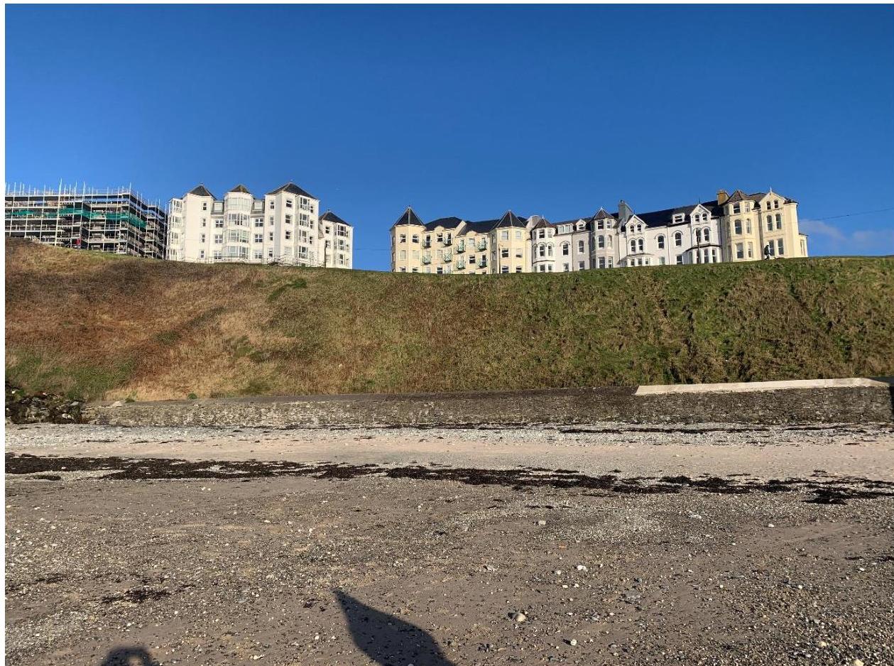 A photograph showing a sandy beach foreground leading up to a grassy embankment, with large white residential or hotel buildings visible on the hilltop in the background.