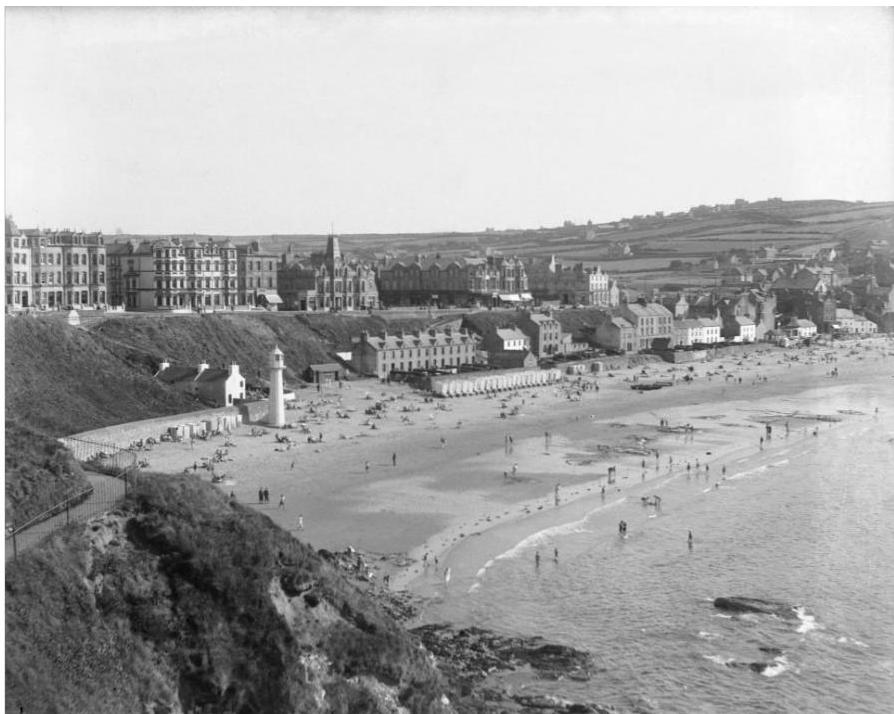 A black and white historical photograph showing a coastal town with a beach, promenade, and rows of beach huts.