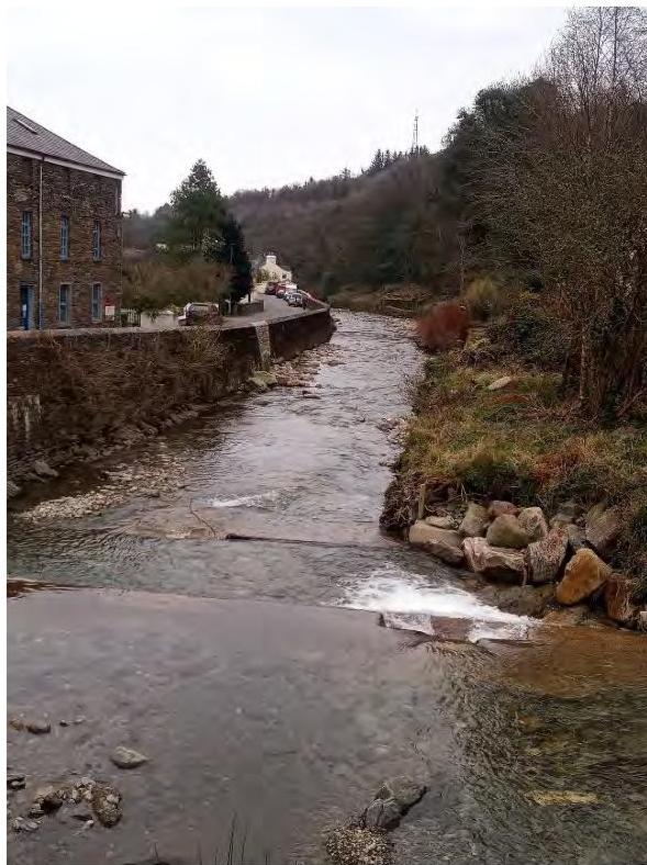 A photograph showing a river flowing through a rural valley with a large stone building on the left bank and a road with parked cars.