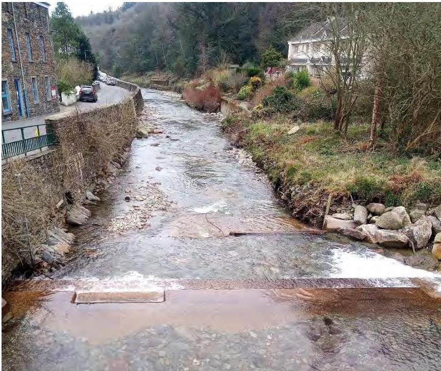 A photograph showing a river flowing through a village with stone buildings on the left and a white house on the right, featuring a weir in the foreground.