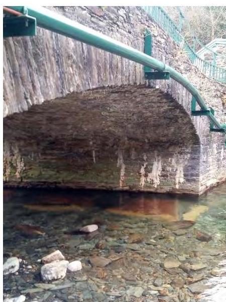 A photograph showing a stone arch structure, likely an aqueduct or bridge, with a green pipe running along the top over a rocky stream bed.