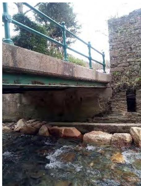 A photograph showing a stone bridge or weir structure with a green metal railing spanning a stream, adjacent to a stone wall.