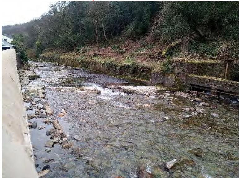 A photograph showing a rocky stream flowing through a wooded area with stone embankments and a partial view of a building structure on the left.