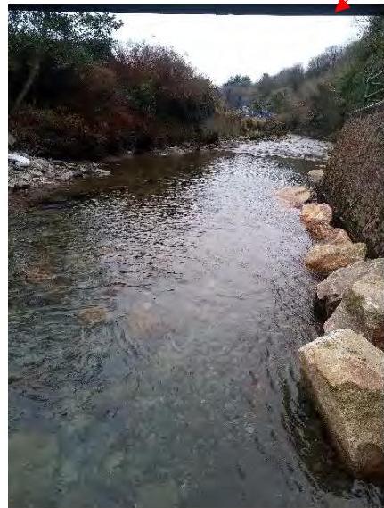 A photograph showing a stream flowing through a wooded area with large rocks lining the right bank, likely for erosion control.