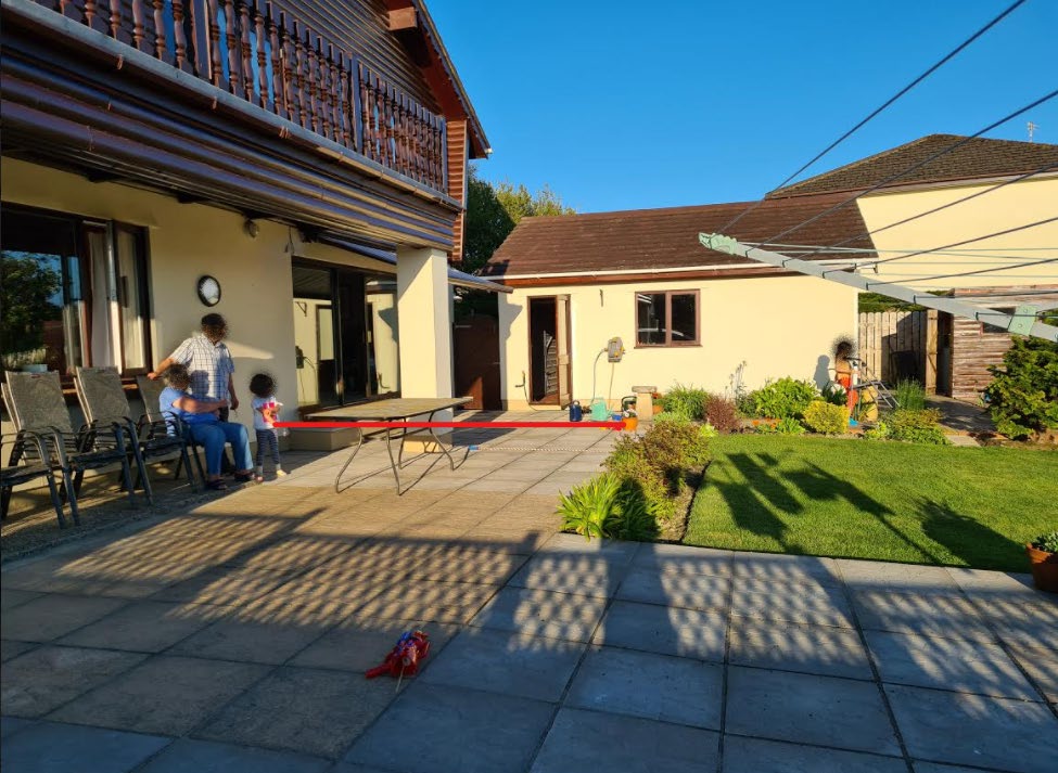 A photograph showing the rear garden and patio area of a residential property, featuring a two-story house with a balcony and a single-story extension or outbuilding.