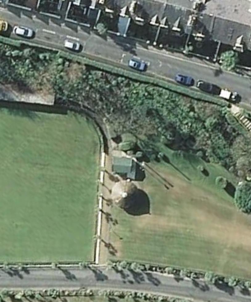 Aerial satellite view showing a green field, a small structure, and a road with parked cars and houses in the background.