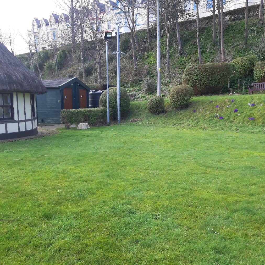 A photograph showing a grassy garden area with a thatched-roof building and a wooden shed on the left, set against a sloping bank with trees and houses in the background.