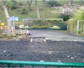 A photograph showing a gravel driveway or parking area leading to a gated entrance with a stone wall and grassy hillside in the background.