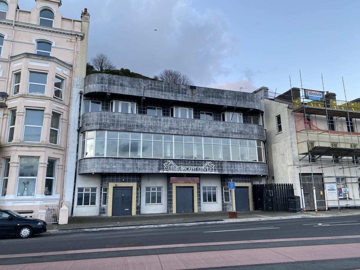 A street-level photograph of a three-story building with a curved grey facade and ground floor commercial units, situated between a cream-colored building and a construction site.