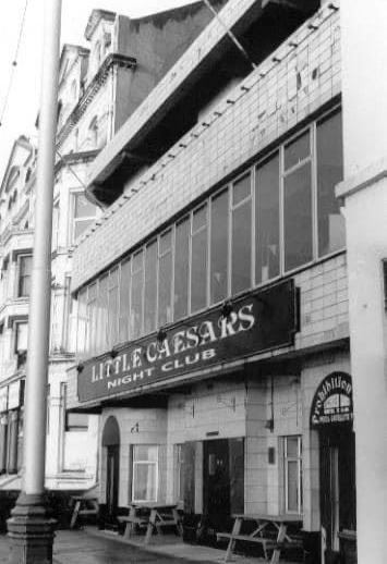 A black and white photograph showing the street-level exterior of a building with a sign reading 'Little Caesars Night Club' and wooden picnic tables on the pavement.