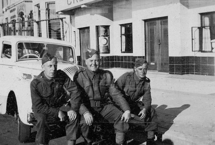A black and white historical photograph showing three men in uniform sitting on the running board of a vintage car in front of a building with a tiled lower facade.