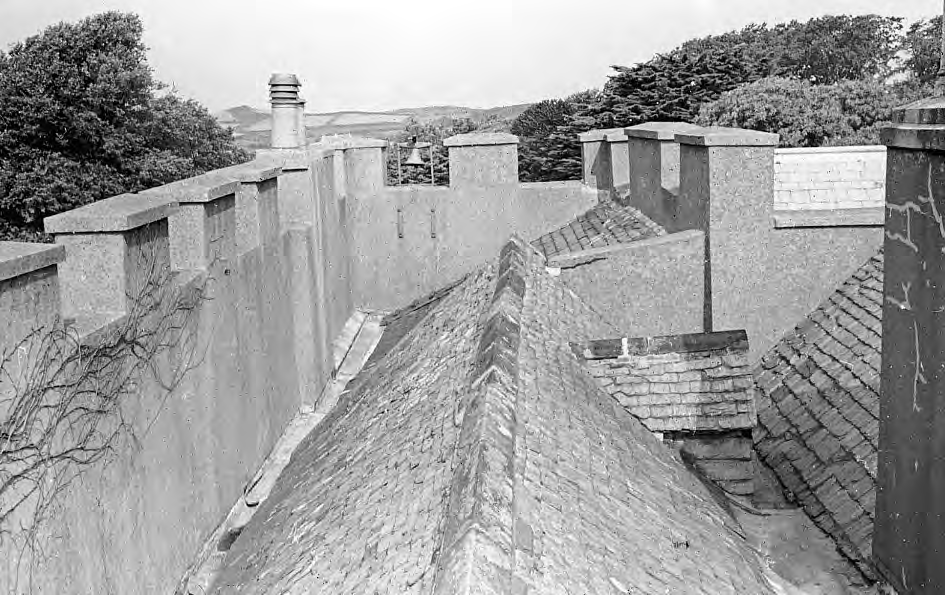 A black and white photograph showing the slate roof, chimneys, and stone parapet of a building in a rural setting.