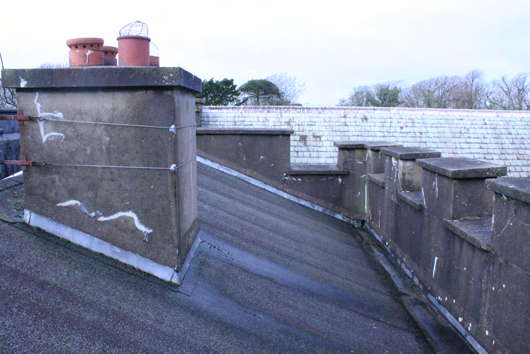 A close-up photograph of a roof section showing a large concrete chimney stack with terracotta pots and a sloping roof surface next to a stepped stone parapet.