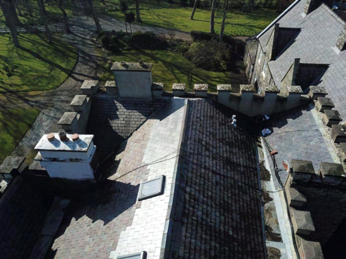 A high-angle photograph showing the slate roof of a historic building with crenellated parapets and chimney stacks. Workers are visible on the roof, indicating ongoing refurbishment works.